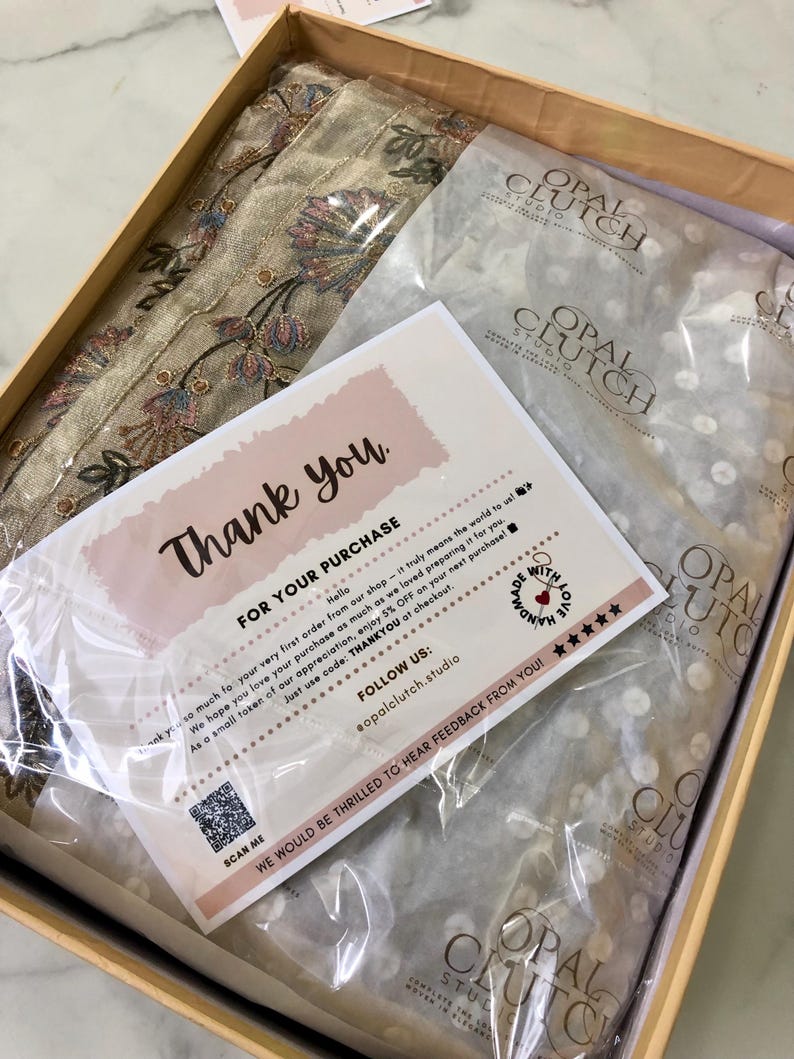May include: A beige box contains a folded textile with floral embroidery, tissue paper with a logo, and a thank you card. The card has text that reads "Thank you" and "For your purchase". The box is on a marble surface.