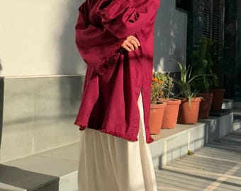 May include: A woman in a burgundy tunic top with white palazzo trousers. The tunic features decorative trim on the sleeves and hem. Gold sandals and earrings complete the outfit. The background shows potted plants and a building, with sunlight casting shadows.