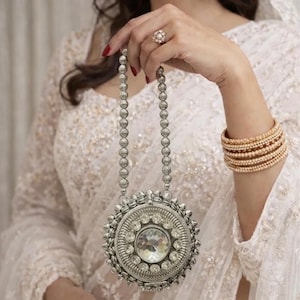 May include: A silver-toned, round handbag with a large, clear central stone and beaded strap. The bag is embellished with smaller stones and a spiky border. The image also shows gold-toned jewelry, including earrings, a ring, and bracelets, and a white, embroidered garment.