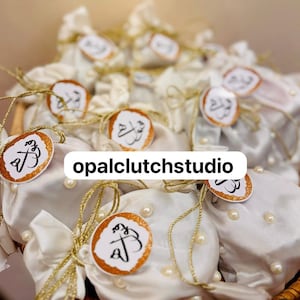 Peut inclure: Plusieurs pochettes en satin blanc, nou&eacute;es avec de la ficelle dor&eacute;e et orn&eacute;es d'&eacute;tiquettes dor&eacute;es avec de la calligraphie. Les pochettes sont d&eacute;cor&eacute;es de fausses perles et dispos&eacute;es dans un panier en osier. Le texte "opalclutchstudio" est visible.