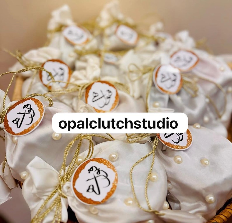 Peut inclure: Plusieurs pochettes en satin blanc, nou&eacute;es avec de la ficelle dor&eacute;e et orn&eacute;es d'&eacute;tiquettes dor&eacute;es avec de la calligraphie. Les pochettes sont d&eacute;cor&eacute;es de fausses perles et dispos&eacute;es dans un panier en osier. Le texte "opalclutchstudio" est visible.