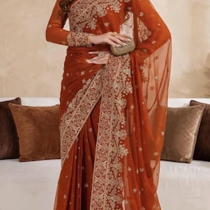 May include: A rust-colored saree featuring intricate gold embroidery along the borders and bodice. The garment includes long sleeves, a sheer fabric overlay, and a matching gold-toned clutch accessory held at the waist, creating a cohesive look.