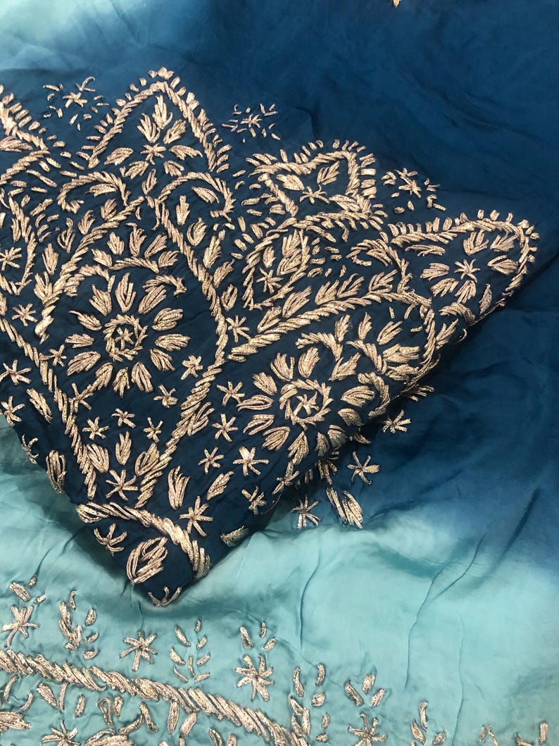 May include: A fabric textile featuring an ombre gradient from deep teal to light turquoise. The fabric is adorned with intricate gold-colored thread embroidery in floral and leaf patterns, concentrated in a dense border design.