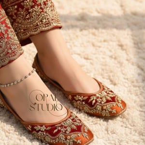 May include: A pair of deep red velvet flat shoes featuring intricate gold embroidery and a matching red velvet pant leg with gold floral patterns. A delicate silver anklet is visible on the ankle. The text OPAL STUDIO is printed in black.
