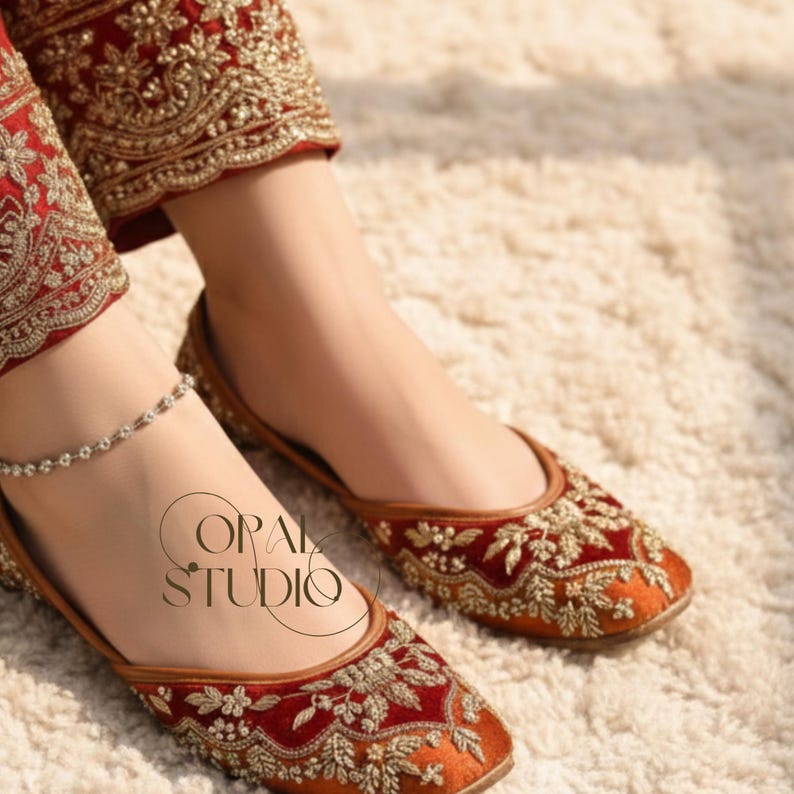 May include: A pair of deep red velvet flat shoes featuring intricate gold embroidery and a matching red velvet pant leg with gold floral patterns. A delicate silver anklet is visible on the ankle. The text OPAL STUDIO is printed in black.