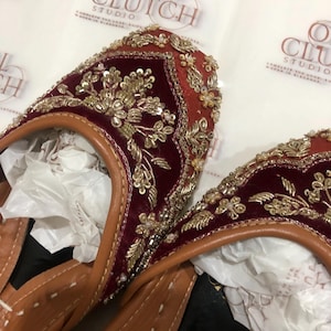 May include: A pair of deep red velvet flats featuring intricate gold embroidery, sequins, and beadwork. The shoes have tan leather trim and white stitching along the edges. The background displays repeating Opal Clutch Studio branding.