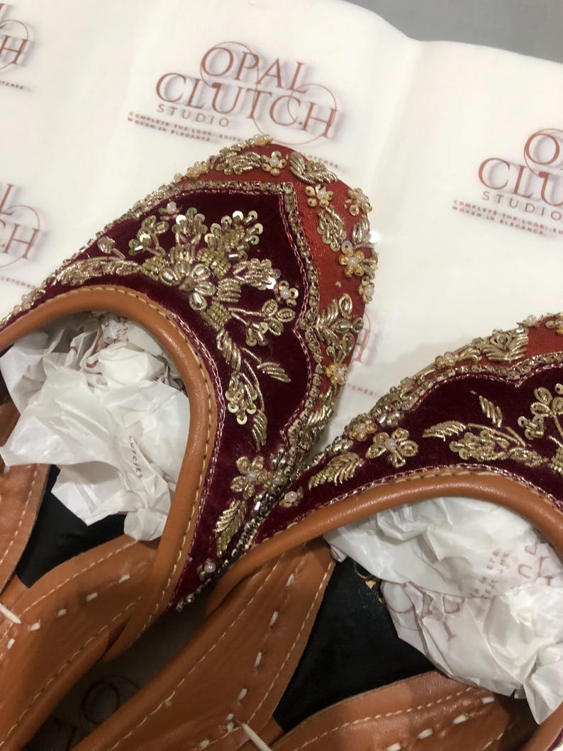 May include: A pair of deep red velvet flats featuring intricate gold embroidery, sequins, and beadwork. The shoes have tan leather trim and white stitching along the edges. The background displays repeating Opal Clutch Studio branding.