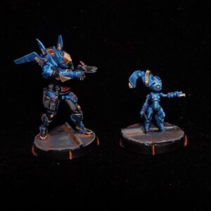 May include: Two miniature figurines of futuristic soldiers. The larger figure is in a blue and black armored suit, holding a weapon. The smaller figure is also in a blue suit, with a unique head design. Both figures stand on circular bases.