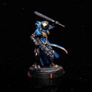 May include: Miniature figurine of a futuristic warrior in blue and gold armor, wielding a sword and a rifle. The figure stands on a circular base with orange accents, set against a dark background. The warrior's helmet has a gold visor.
