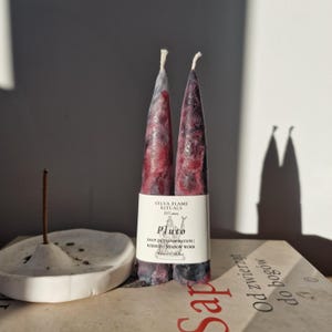 May include: Two maroon taper candles with a marbled effect, labelled "Pluto". A white ceramic incense holder with an incense stick is next to the candles. The candles are on a book. The candles are a decorative item.