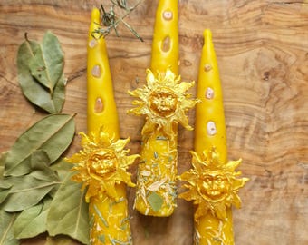 Puede incluir: Tres velas cónicas amarillas decoradas con emblemas de sol dorados, pequeñas piedras claras incrustadas y hierbas secas esparcidas. Las velas están dispuestas verticalmente y presentan una superficie texturizada con inclusiones botánicas.