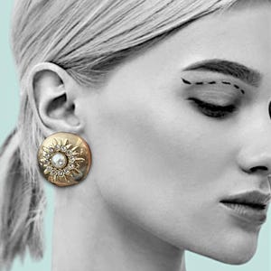 May include: A gold-toned, round earring with a central pearl surrounded by small, clear crystals. The earring has a sunburst design and is set against a grayscale image of a person's face.