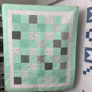 May include: A handmade quilt in shades of mint green, white, and gray. The quilt features a grid pattern of squares with various designs, including stars and polka dots. The quilt has a white border and a mint green edge.