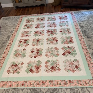 May include: A large, rectangular quilt with a floral pattern. The quilt features a white background with repeating blocks of red, green, and pink floral fabric. A light green border surrounds the central design, and a floral patterned border frames the quilt.