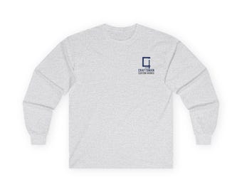 May include: A light gray long-sleeve shirt with a crew neck. The shirt features a small navy blue square logo with the text "CRAFTSMAN CUSTOM WORKS" printed below it. The shirt is made of a soft, comfortable material.