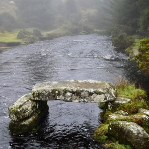 Puede incluir: Un puente de piedra sobre un río en un bosque brumoso. El puente está hecho de grandes rocas cubiertas de musgo. El río es oscuro y fluye. El bosque es denso y verde.