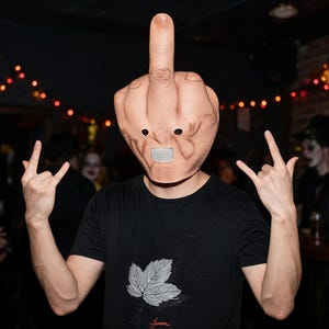 May include: A full-head mask shaped like a hand making a middle finger gesture, featuring skin-tone coloring, two circular eye holes, and a rectangular mesh mouth opening, worn with a black t-shirt displaying a white leaf graphic on the chest.