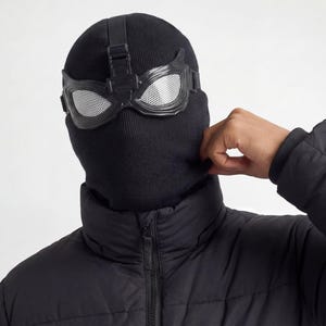 May include: A black knitted balaclava with attached black framed goggles with a silver mesh lens. The balaclava is worn with a black puffer jacket. The person is adjusting the balaclava.