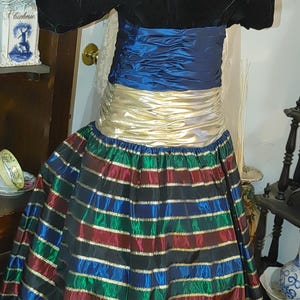 May include: A vintage formal dress with a black velvet bodice and puffed sleeves. The waist is cinched with a blue and gold sash, and the skirt features horizontal stripes in red, green, blue, and gold. The dress is displayed on a mannequin.