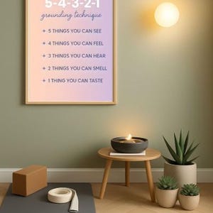 May include: A framed poster with the text "5-4-3-2-1 grounding technique" hangs on a green wall. The poster is above a yoga mat, block, and tape. A small table holds a candle and a plant. A light fixture is on the wall.