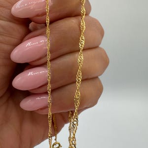 May include: A gold-coloured chain necklace with a twisted rope design. The necklace is held up against a white background, showcasing its intricate details and the warm tone of the metal. The chain is a fine piece of jewellery.