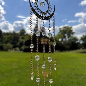 May include: A gold-toned metal sun and crescent moon wall hanging featuring multiple dangling chains. The piece is adorned with clear, faceted crystal beads and small circular sunburst charms, with a larger crystal suspended in the center.