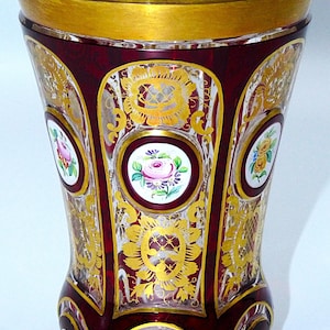 May include: A deep red glass vase featuring a wide gold-painted rim and intricate gold filigree patterns. The exterior is decorated with multiple circular white medallions, each containing a hand-painted floral motif in various colors.