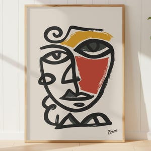Peut inclure: Impression d'art abstrait représentant un visage avec des lignes noires audacieuses, une joue rouge et des touches de jaune. L'œuvre, signée "Picasso", est encadrée dans un cadre en bois clair et exposée sur un mur blanc.