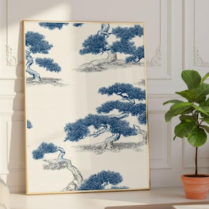 May include: A framed art print featuring a repeating pattern of stylized blue bonsai trees on a cream background. The artwork is displayed in a gold frame and leans against a white wall. A potted plant and a coffee table are in the foreground.