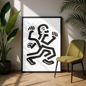 Puede incluir: Impresi&oacute;n art&iacute;stica abstracta en blanco y negro de una figura con brazos y piernas extendidos. La obra de arte est&aacute; enmarcada en un marco negro y se muestra sobre un fondo blanco. Una silla verde y plantas en macetas est&aacute;n en el fondo.