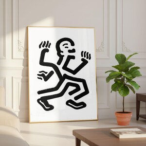 Puede incluir: Impresi&oacute;n art&iacute;stica abstracta en blanco y negro que representa una figura estilizada con m&uacute;ltiples extremidades en movimiento. La obra de arte est&aacute; enmarcada en un marco dorado y se apoya contra una pared blanca. Una planta en maceta y una mesa de madera est&aacute;n en primer plano.