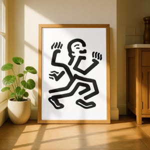 Puede incluir: Una impresi&oacute;n art&iacute;stica enmarcada que presenta una figura abstracta en blanco y negro en movimiento. La obra de arte, con sus l&iacute;neas audaces y estilo minimalista, se encuentra sobre un fondo blanco dentro de un marco de madera natural. La impresi&oacute;n se exhibe sobre un suelo de madera.