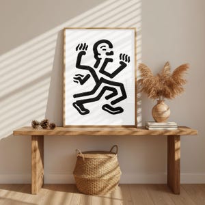 Puede incluir: Una impresi&oacute;n art&iacute;stica enmarcada que presenta una figura abstracta en blanco y negro con m&uacute;ltiples extremidades. La obra de arte se exhibe en un banco de madera con una cesta tejida, un jarr&oacute;n y plantas secas. La habitaci&oacute;n tiene una paleta de colores neutros.