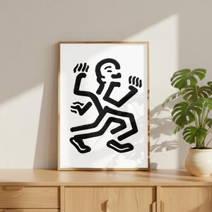 Puede incluir: Una impresi&oacute;n art&iacute;stica enmarcada que presenta una figura abstracta en blanco y negro en movimiento. La obra de arte representa una persona estilizada con m&uacute;ltiples brazos y piernas, sobre un fondo blanco. El marco es de color marr&oacute;n claro y la impresi&oacute;n se exhibe en un mueble de madera.