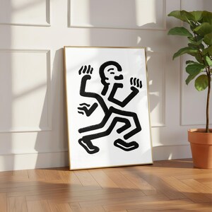 Puede incluir: Una impresi&oacute;n art&iacute;stica enmarcada con un marco dorado, que representa una figura abstracta en blanco y negro en movimiento. La obra de arte utiliza l&iacute;neas gruesas y audaces y un dise&ntilde;o minimalista. La impresi&oacute;n est&aacute; apoyada contra una pared blanca sobre un suelo de madera.