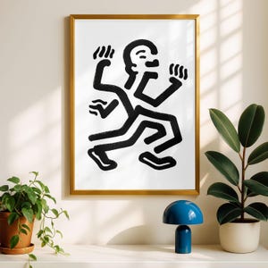 Puede incluir: Una impresi&oacute;n art&iacute;stica enmarcada con una figura abstracta en blanco y negro en movimiento. La obra de arte est&aacute; en un marco dorado y cuelga de una pared blanca. Una l&aacute;mpara azul y plantas en macetas est&aacute;n en un estante blanco.