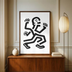 Puede incluir: Una impresi&oacute;n art&iacute;stica abstracta en blanco y negro enmarcada que representa una figura estilizada en movimiento. La obra de arte tiene un dise&ntilde;o minimalista con l&iacute;neas negras gruesas sobre un fondo blanco. La impresi&oacute;n se exhibe en un mueble de madera con una l&aacute;mpara y un jarr&oacute;n.