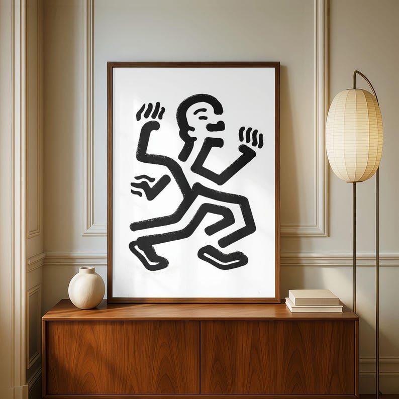 Puede incluir: Una impresi&oacute;n art&iacute;stica abstracta en blanco y negro enmarcada que representa una figura estilizada en movimiento. La obra de arte tiene un dise&ntilde;o minimalista con l&iacute;neas negras gruesas sobre un fondo blanco. La impresi&oacute;n se exhibe en un mueble de madera con una l&aacute;mpara y un jarr&oacute;n.