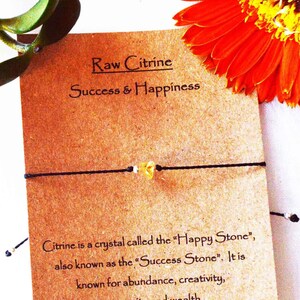 May include: A brown card with the text "Raw Citrine Success & Happiness" and a black string bracelet with a small yellow citrine crystal. The card also says "Citrine is a crystal called the "Happy Stone", also known as the "Success Stone". It is known for abundance, creativity, prosperity and wealth. Adjustable sliding knot."