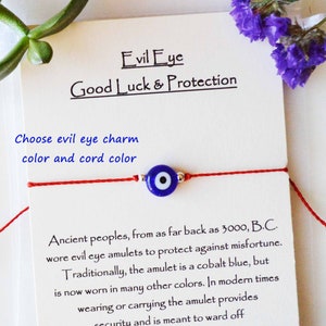 May include: A red cord bracelet featuring a blue evil eye charm with silver accents. The card behind the bracelet reads "Evil Eye Good Luck & Protection" and includes text about the amulet's history and purpose.