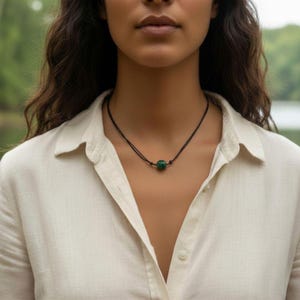 May include: A minimalist necklace featuring a dark cord and a vibrant green, cylindrical bead. The necklace is worn against a light-colored button-down shirt, creating a contrast that highlights the jewelry's design.