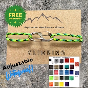 May include: A climbing bracelet with a green and black cord, connected by a silver carabiner. The card behind the bracelet has the words "Climbing", "Exploration, Resilience, Altitude", and "Adjustable Waterproof!"