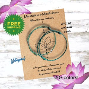 May include: Two teal-colored string bracelets with a lotus flower design on a brown card. The card reads "Meditation & Mindfulness" and "Wear this as a reminder." Additional text includes "With or without bead," "Waterproof," and "20+ colors!"