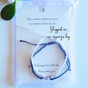 May include: Two friendship bracelets, one white and one navy blue, are displayed inside a clear organza bag. The bracelets are knotted and have a small silver charm. A card with the text "Us" and a message is visible.
