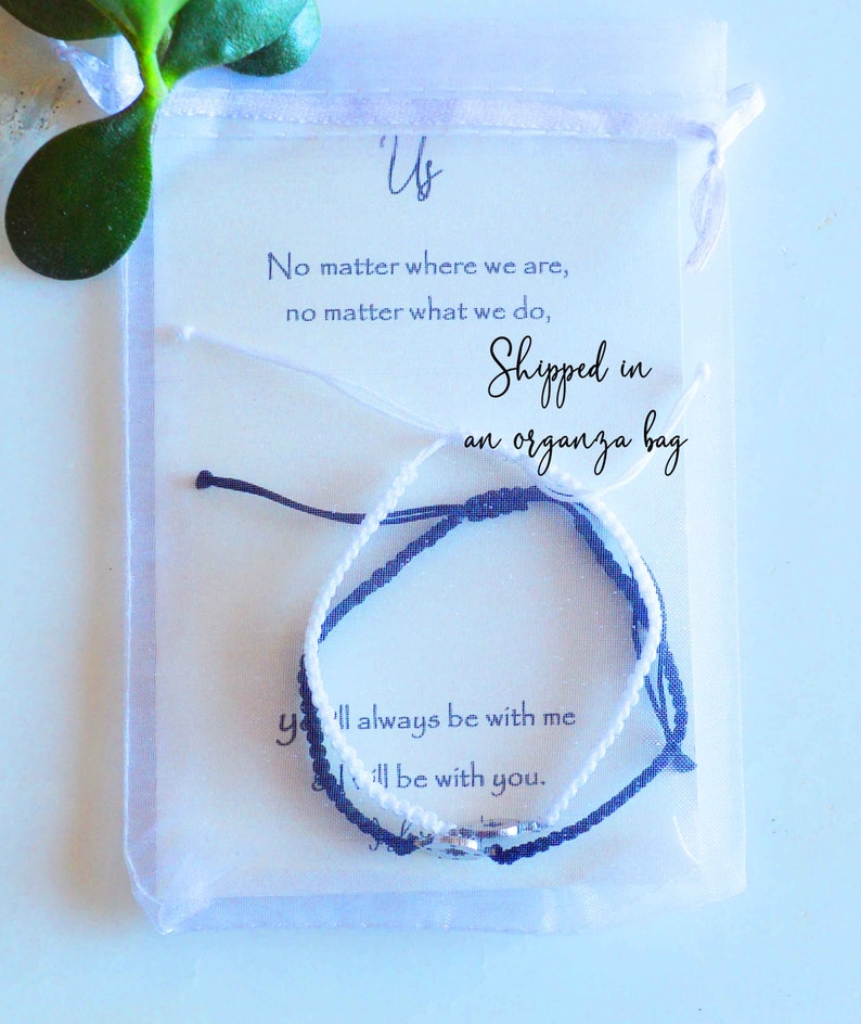 May include: Two friendship bracelets, one white and one navy blue, are displayed inside a clear organza bag. The bracelets are knotted and have a small silver charm. A card with the text "Us" and a message is visible.