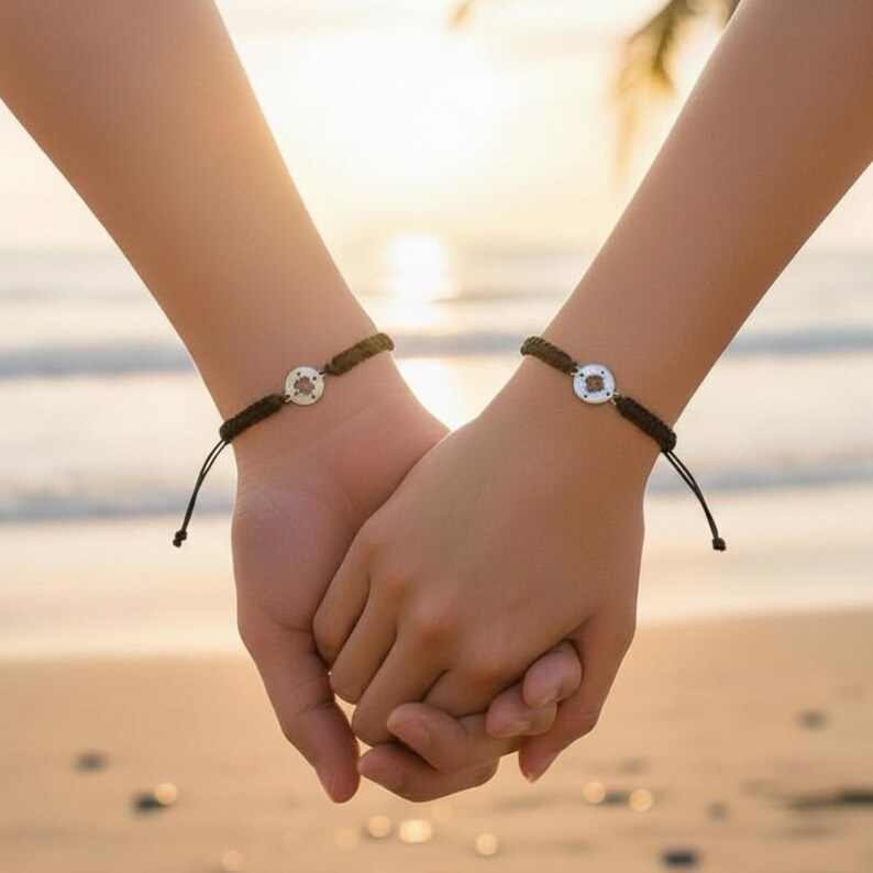 May include: Two black cord bracelets with a circular silver charm are worn on wrists. The bracelets are tied with adjustable knots. The hands are clasped together, suggesting a connection or relationship. The background is a blurred beach scene.