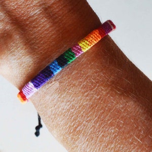 May include: A rainbow colored friendship bracelet with a black cord tie. The bracelet is made of woven thread and is worn on a person's wrist.