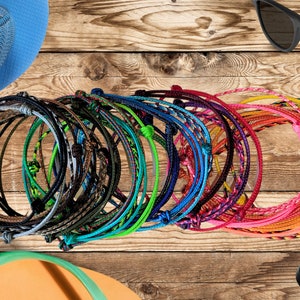 May include: A collection of colorful braided bracelets in various shades of blue, green, red, pink, and yellow. The bracelets are made of woven string and have a knotted closure.