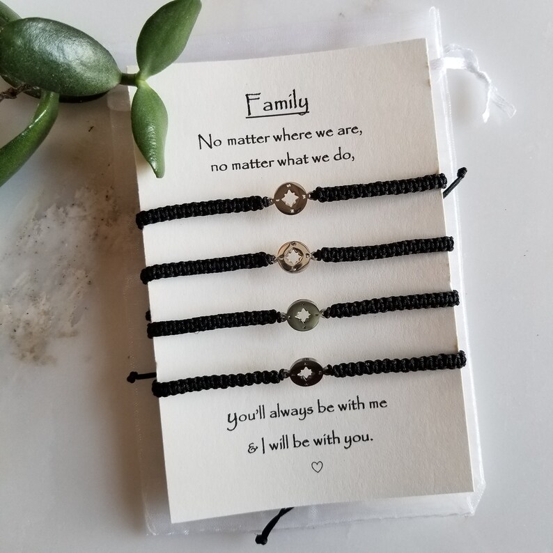 May include: Four black woven bracelets with silver compass charms are displayed on a white card. The card reads "Family" and includes a sentimental quote. The bracelets are presented in a small, clear bag.