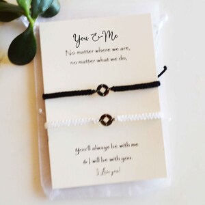 May include: A pair of matching black and white friendship bracelets with a compass charm, displayed on a card with the text "You & Me". The card also includes the phrases "No matter where we are" and "You'll always be with me".